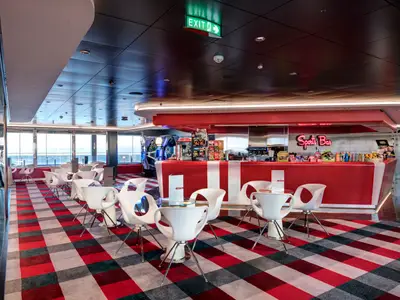 Blick in die Sports Bar an Bord der MSC Meraviglia. Der Raum zeigt eine rot-weiß gestreifte Theke, weiße Stühle und Tische auf einem karierten Teppich und große Fenster mit Meerblick.