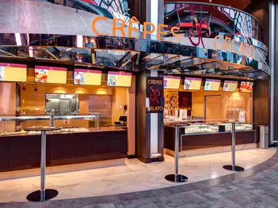 Innenansicht des Jean-Philippe Crêpes & Gelato an Bord der MSC Meraviglia mit Verkaufstheken, einer Vielzahl an Süßspeisen und digitalen Menütafeln. Die Decke ist verspiegelt, und ein glänzender Boden führt durch den Bereich. Keine Personen sichtbar.