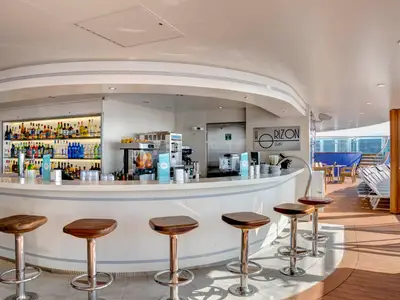 Die moderne, geschwungene Horizon Bar an Bord der MSC Meraviglia mit zahlreichen Spirituosen, Barkeeper-Bereich und Barhockern. Im Hintergrund sind Sonnenliegen und das Meer zu sehen.