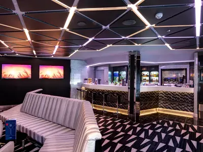Die stylische TV Studio & Bar an Bord der MSC Meraviglia mit einem langen Bartresen, bequemen Lounge-Sitzmöbeln und auffälliger dreieckiger Deckenbeleuchtung. Zwei Bildschirme zeigen Sonnenuntergänge.