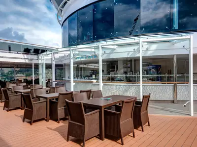 Die Atmosphere Ice Cream Bar an Bord der MSC Meraviglia. Das Deck ist mit braunen Rattan-Sitzgruppen ausgestattet. Im Hintergrund die Bar und eine städtische Skyline unter bewölktem Himmel.