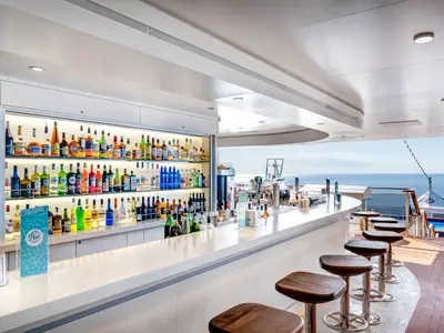 Die Horizon Bar an Bord der MSC Meraviglia mit umfangreicher Auswahl an Spirituosen. Moderne Barhocker. Im Hintergrund weites Meer und blauer Himmel. Eine einladende Outdoor-Bar-Szene.