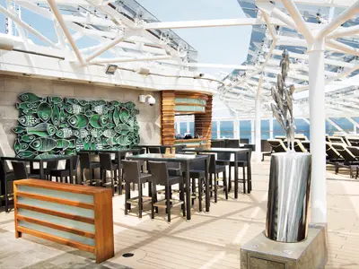 Eine moderne Outdoor-Bar mit Hochtischen und stilvoller Fisch-Wandkunst auf dem Sonnendeck der MSC Meraviglia. Liegestühle und das offene Meer sind im Hintergrund sichtbar.