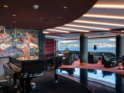 Die luxuriöse Top Sail Lounge an Bord der MSC Meraviglia zeigt einen schwarzen Flügel, gemütliche Sessel, eine farbenfrohe Kunstwand und grosse Fenster mit Blick auf die Küste und das Meer.