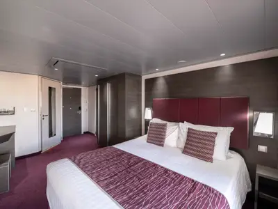 Moderne, barrierefreie Kabine an Bord der MSC Meraviglia. Doppelbett mit lila Tagesdecke. Links Schreibtisch und Schränke. Lilafarbener Teppich und Holzelemente.