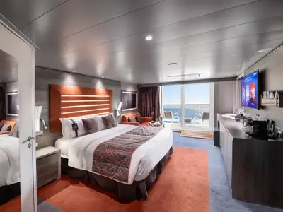 Helle MSC Yacht Club Deluxe Suite an Bord der MSC Meraviglia. Man sieht ein großes Bett, ein Sofa, ein Sideboard mit TV und Kaffeemaschine sowie einen Balkon mit Meerblick.