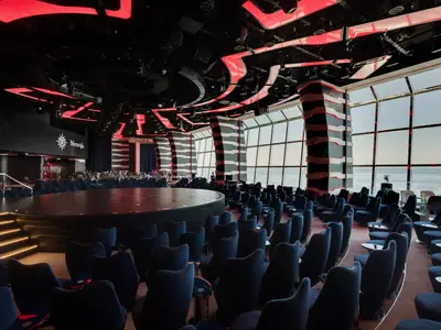 Blick in die moderne Carousel Lounge der MSC Meraviglia mit runder Bühne, blauen Sesseln und grossen Fenstern zum Meer. Rote Lichtakzente.