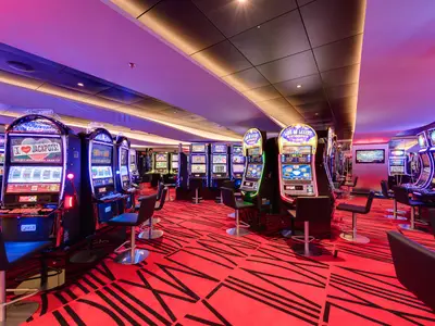 Casino Imperiale der MSC Meraviglia. Viele leuchtende Spielautomaten und Barhocker auf rotem Teppich. Elegante Deckenbeleuchtung.