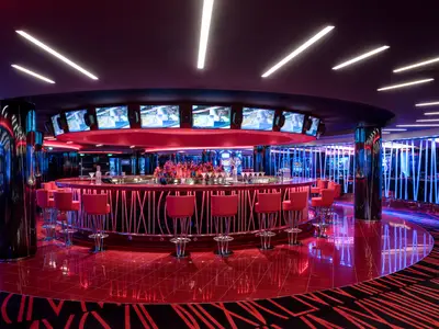 Kreisrunde, moderne Bar mit roter Theke und Barhockern im Casino Imperiale an Bord der MSC Meraviglia. Über dem Tresen sind Bildschirme mit Videos angebracht. Der glänzende rote Boden reflektiert das bunte Neonlicht, das eine lebhafte Atmosphäre schafft. Eine grosse Auswahl an Getraenken steht bereit. Keine Personen.