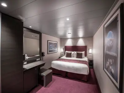 Eine moderne Innenkabine an Bord der MSC Meraviglia. Zu sehen sind ein grosses Doppelbett, ein Schminktisch mit Spiegel und ein Kleiderschrank. Der Raum ist in dunklen Holztoenen und Beerenfarben gehalten.