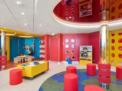 Ein lebhafter, bunter LEGO-Kinderbereich an Bord der MSC Meraviglia mit Spieltischen, Hockern und farbenfrohen Wänden. Ein großes LEGO-Männchen ziert eine blaue Wand.