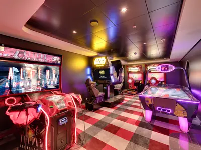 An interior view of the Virtual Games Arcade on the MSC Meraviglia cruise ship. The brightly lit room features several arcade machines, including a red 'Aliens Armageddon' game on the left, a 'Star Wars Battle Pod' in the center, and an air hockey table with purple lighting on the right. The checkered floor is red, black, and grey. No people are visible.