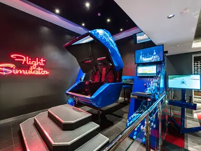 Ein blauer Flugsimulator mit zwei Sitzen und einer leuchtenden roten Neon-Schrift 'Flight Simulator' an einer dunklen Wand an Bord der MSC Meraviglia. Mehrere Bildschirme zeigen Flugsimulationen.