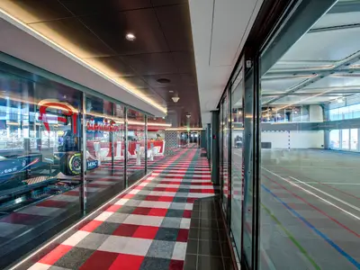 Gang an Bord der MSC Meraviglia mit F1-Rennsimulator hinter Glas links und Indoor-Sportplatz rechts. Rot-schwarz-grauer Teppich im Gang, Tageslicht.
