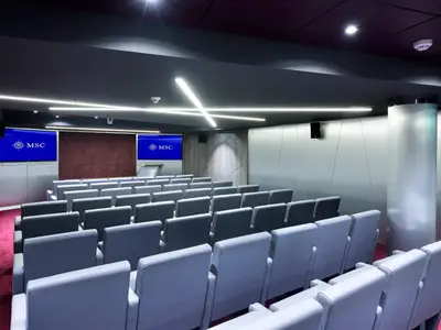 Moderner Konferenzraum der MSC Meraviglia. Graue Reihenbestuhlung, roter Teppich, zwei blaue MSC-Bildschirme und LED-Deckenbeleuchtung sind sichtbar.