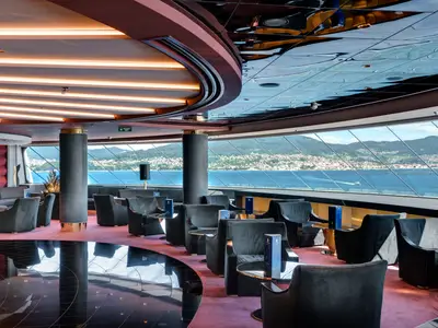 Eine elegante Lounge der MSC Meraviglia mit bequemen Sesseln, dunklem Teppich und großen Fenstern mit Panoramablick auf die Küste. Decke mit Lichtstreifen.