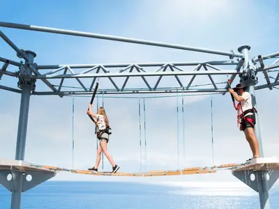 Eine junge Frau und ein Mann ueberqueren die Himalayan Bridge, einen Hochseilgarten auf Deck der MSC Meraviglia, ueber dem blauen Meer unter sonnigem Himmel.
