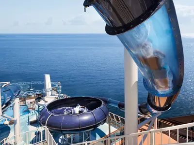 Eine spektakuläre blaue Wasserrutsche an Bord der MSC Meraviglia. Man sieht den Trichter und Teile des Rutschenrohrs. Im Hintergrund erstreckt sich das weite, glitzernde Meer unter klarem Himmel.