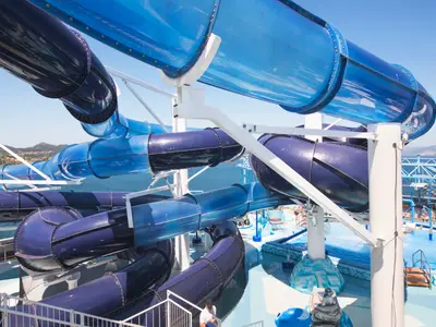 Gewundene blaue Wasserrutschen des Polar Aquaparks auf dem Deck der MSC Meraviglia. Meer und Küste im Hintergrund. Einige Badegäste am Pool sichtbar.