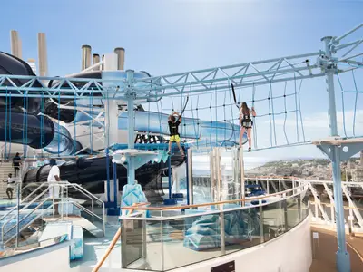 Oberdeck der MSC Meraviglia mit Polar Aquapark. Grosse Wasserrutschen und die Himalayan Bridge mit zwei kletternden Personen. Im Hintergrund Meer und Küstenstadt unter blauem Himmel.