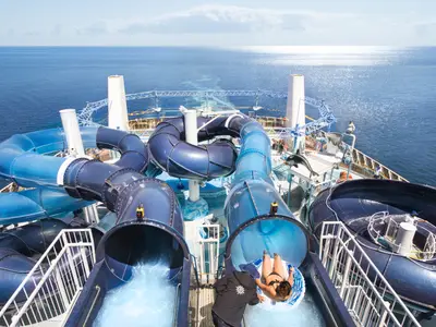 Vogelperspektive auf den Polar Aquapark an Bord der MSC Meraviglia. Blaue Wasserrutschen mit Badegästen und Crew auf dem Deck. Im sonnigen Hintergrund glitzert das weite Meer.