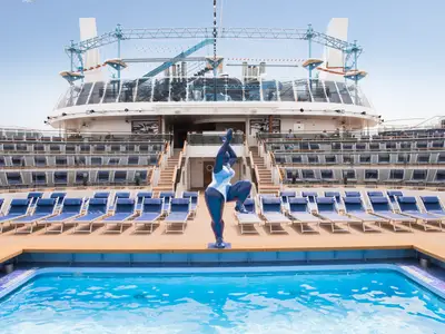 Weitwinkelansicht des Horizon Pools und Amphitheaters an Bord der MSC Meraviglia. Blaue Sonnenliegen und eine moderne Skulptur vor der gestuften Tribüne.