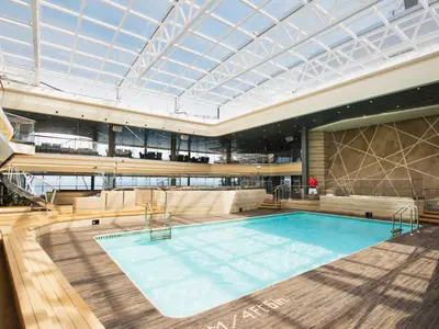 Blick auf den hellen Indoor-Poolbereich "Bamboo Pool" der MSC Meraviglia. Tageslicht fällt durch das transparente Glasdach auf das türkisfarbene Wasser und das umliegende Holzdeck. Im Hintergrund sind Sitzbereiche und der weite Meerblick durch Panoramafenster zu erkennen. Ein Rettungsschwimmer steht am Beckenrand.
