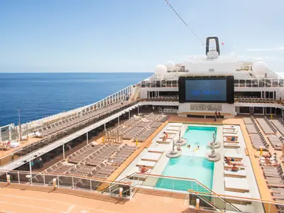 Blick von oben auf das lebhafte Sonnendeck der MSC Meraviglia: Der große Atmosphere Pool, zahlreiche Sonnenliegen und ein riesiger LED-Bildschirm. Im Hintergrund das weite, blaue Meer.