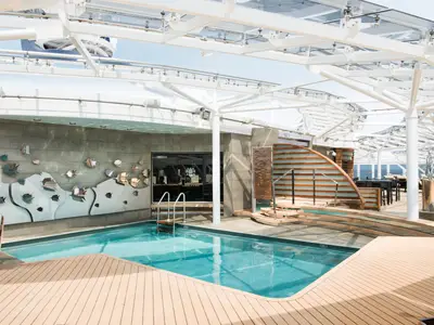 MSC Meraviglia: Der überdachte Poolbereich des MSC Yacht Clubs. Blauer Pool, Holzdecks und Fisch-Kunst an der Wand. Eine Bar sowie Lounge-Möbel sind sichtbar.