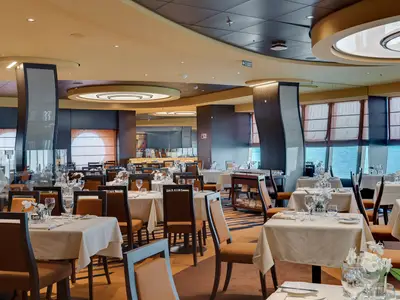 Elegantes Panoramarestaurant an Bord der MSC Meraviglia. Viele gedeckte Tische mit weißen Tischdecken und braunen Stühlen. Große Fensterfronten mit Blick auf das weite, blaue Meer.
