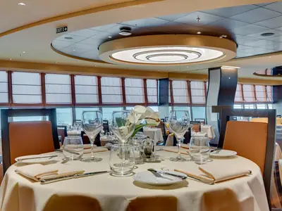 Blick ins Panorama Restaurant der MSC Meraviglia. Ein elegant gedeckter Tisch mit Weingläsern, Geschirr und einer Orchidee ist im Vordergrund. Fenster geben den Blick auf das Meer frei.