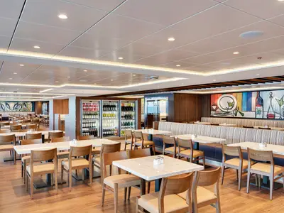 Helles, geräumiges Marketplace Buffet Restaurant an Bord der MSC Meraviglia mit Holzböden, weissen Marmortischen, Holzsesseln. Gekühlte Getränke und bunte Wandkunst zieren den Raum.