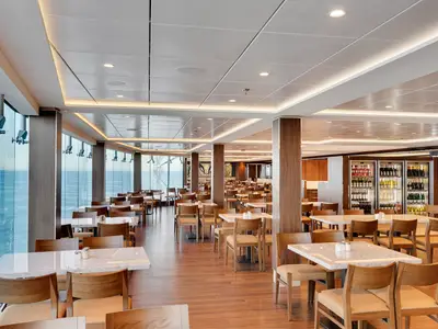 Das geräumige Marketplace Buffet Restaurant an Bord der MSC Meraviglia bietet helle Holzböden, Marmortische und große Fenster mit weitem Blick auf das Meer. Im Hintergrund sind gekühlte Getränkeauslagen sichtbar.