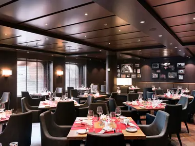 Innenansicht des Spezialitätenrestaurants Butcher's Cut an Bord der MSC Meraviglia. Elegante Einrichtung mit dunklem Holz, roten Tischsets und Jalousienfenstern. Warme Beleuchtung. Ohne Gäste.