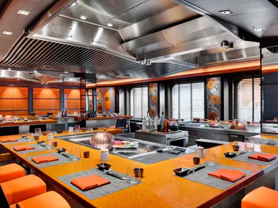 Modernes Kaito Teppanyaki Restaurant der MSC Meraviglia. Orangefarbene Tische mit Gedecken umgeben die Kochstationen. Elegante Metall-Decken und Fenster mit Jalousien prägen das Innere.