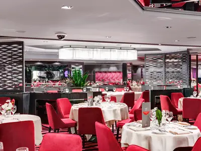 Geräumiges, modernes Restaurant an Bord der MSC Meraviglia mit vielen runden Tischen, eingedeckt mit weißen Tischtüchern und Gläsern. Die Sessel sind in leuchtendem Rot gehalten. Dekorative Trennwände und eine große, längliche Lichtinstallation zieren den Raum.
