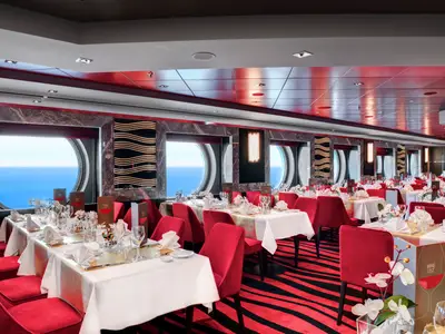 Das helle Waves Restaurant an Bord der MSC Meraviglia mit runden Bullaugenfenstern zum Meer. Rote Stühle und weiß gedeckte Tische mit goldenen Läufern sind für Gäste vorbereitet.