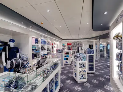 Heller, moderner MSC Shop an Bord der MSC Meraviglia. Weiße Regale zeigen Kleidung, Mützen, Taschen, Plüschtiere, Gadgets. Mannequins tragen MSC-Outfits. Teppichboden mit maritimem Sternenmuster.