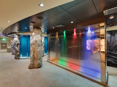 Ein moderner Spa-Bereich an Bord der MSC Meraviglia mit Gesteins-Säulen, blauen Glasduschen und einer großen Glaswand mit farbigen Lichtduschen in Rot, Grün und Blau.