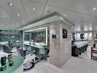Ein moderner Barbershop an Bord der MSC Meraviglia mit Friseurstühlen, Waschbecken, großen Spiegeln und Regalen voller Pflegeprodukte. Die helle Einrichtung ist in Grün- und Cremetönen gehalten.