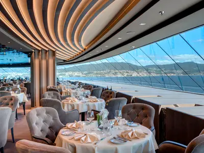 Elegantes Restaurant an Bord der MSC Meraviglia. Gedeckte Tische, Samtsessel. Panoramafenster mit Blick auf Küstenlandschaft, Meer und Berge.