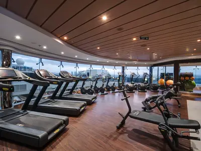 Heller Fitnessraum der MSC Meraviglia mit Technogym-Laufbändern und Ergometern. Große Panoramafenster bieten Meerblick und Sicht auf das Außendeck. Holzboden und helle Decke. Ohne Personen.