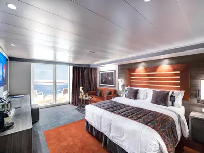 Blick in eine helle und moderne MSC Yacht Club Deluxe Suite an Bord der MSC Meraviglia. Ein großes Doppelbett, ein Sofa, ein Flachbildfernseher und ein Balkon mit Meerblick sind zu sehen.