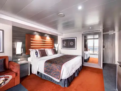 Geräumige MSC Yacht Club Deluxe Suite an Bord der MSC Meraviglia. Das Bild zeigt ein Doppelbett, ein Sofa, einen Nachttisch und einen Spiegel, der den Balkon reflektiert.