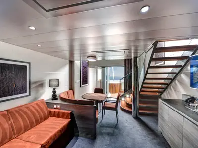 Ansicht der stilvollen MSC Yacht Club Duplex Suite an Bord der MSC Meraviglia mit oranger Couch, Esstisch, Treppe zum Obergeschoss und Zugang zum Balkon mit Whirlpool.