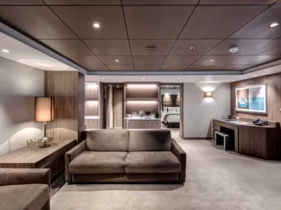 Eine moderne, luxuriöse Suite an Bord der MSC Meraviglia. Im Vordergrund ein Wohnbereich mit braunem Sofa. Im Hintergrund ist ein Schlafzimmer mit Doppelbett und ein Barbereich sichtbar.