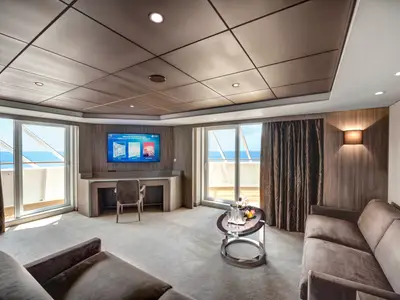 Blick in eine geräumige, luxuriöse Suite der MSC Meraviglia. Sie verfügt über einen eleganten Wohnbereich mit Sofa, Schreibtisch, Couchtisch und zwei großen Balkontüren, die den Blick auf das weite Meer freigeben. Auf dem Couchtisch stehen Champagner und Früchte.