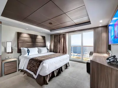 Geräumige Royal Suite der MSC Meraviglia. Doppelbett, Nachttisch, Schreibtisch, Wand-TV. Glastüren zum Balkon mit Meerblick. Moderne Einrichtung mit Holzakzenten.