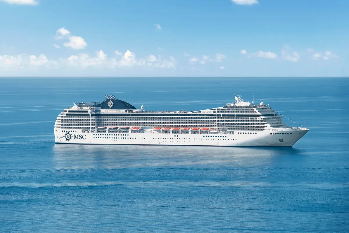 Die MSC Musica, ein weißes Kreuzfahrtschiff, gleitet auf blauem, ruhigem Meer unter einem blauen Himmel mit weißen Wolken. Das Schiff ist in Seitenansicht von oben fotografiert, mit dem markanten MSC-Logo am Heck.