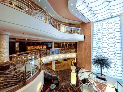 Das helle, mehrstöckige Atrium der MSC Musica. Eine elegante, geschwungene Treppe mit Glas- und Goldgeländer ist zentral. Rechts ein hohes Fenster mit blauem Schuppenmuster und einer Palme. Boutiquen säumen die oberen Ebenen.